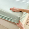 A hand is folding a light mint green fitted bed sheet on a white mattress.