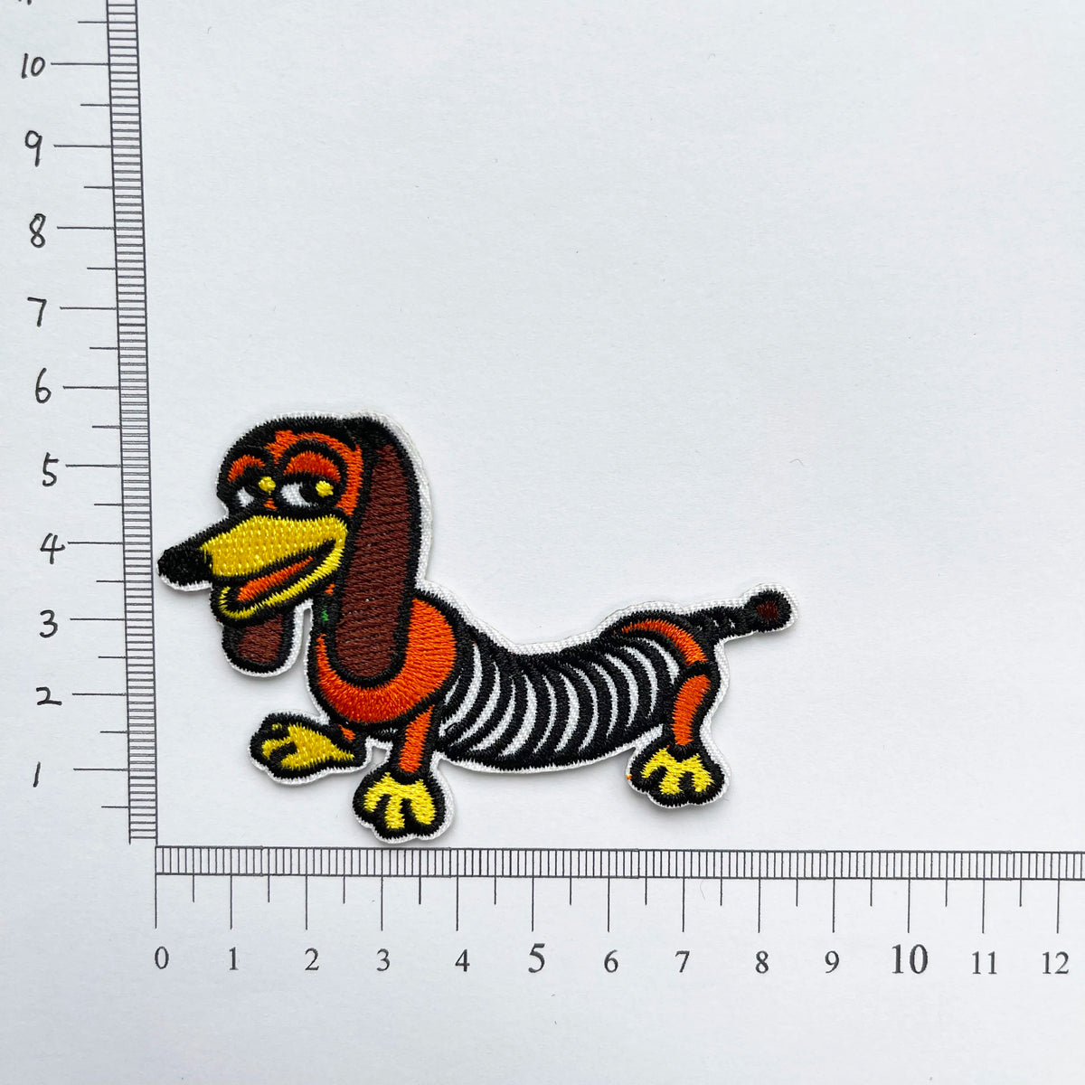 An embroidered patch of a dog with long brown ears and an orange body, shown against a white ruler.
