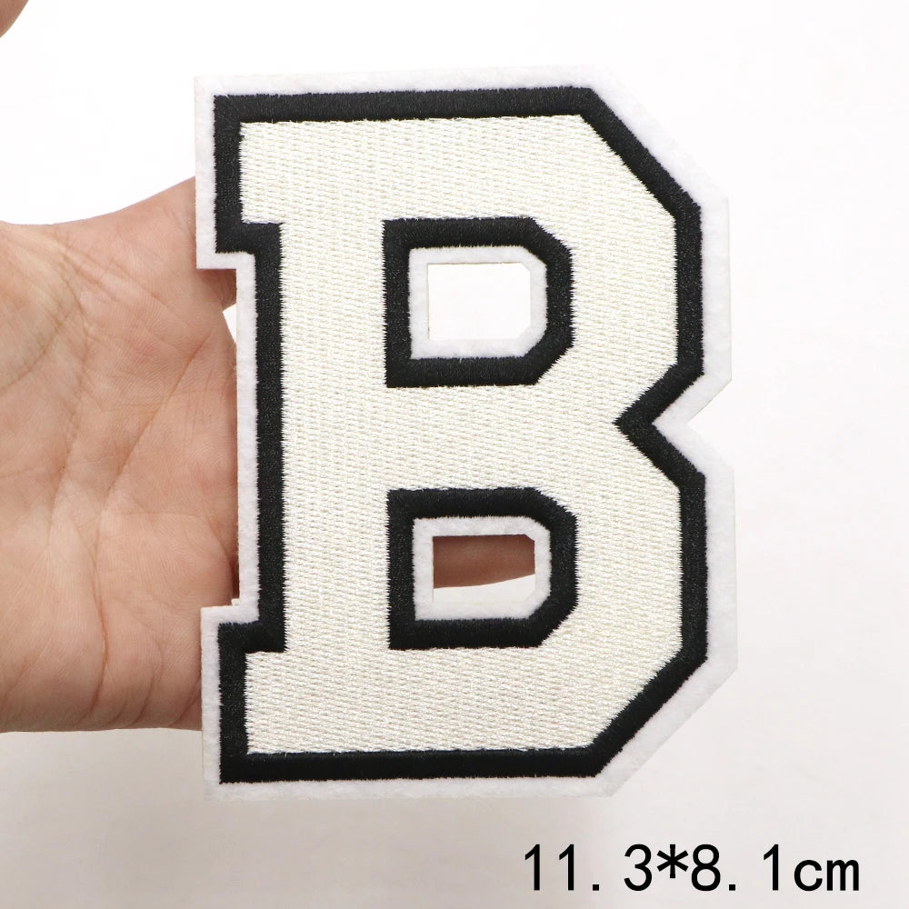 A hand holds a white letter B patch with black outline and embroidery on a white background.