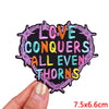 A hand holds a colorful embroidered patch with the text Love Conquers All Even Thorns.