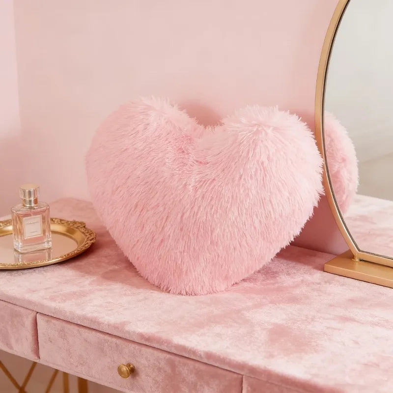 A pink heart-shaped plush cushion resting on a pink vanity table next to a gold mirror.