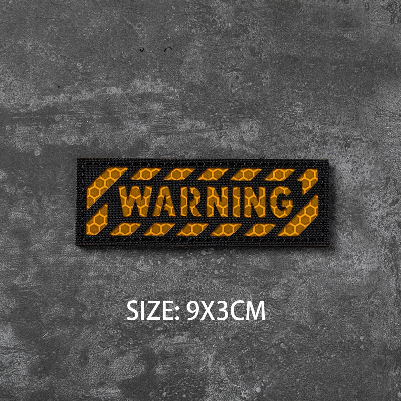 A black embroidered warning badge with yellow text on a dark gray textured background, size 9x3cm.
