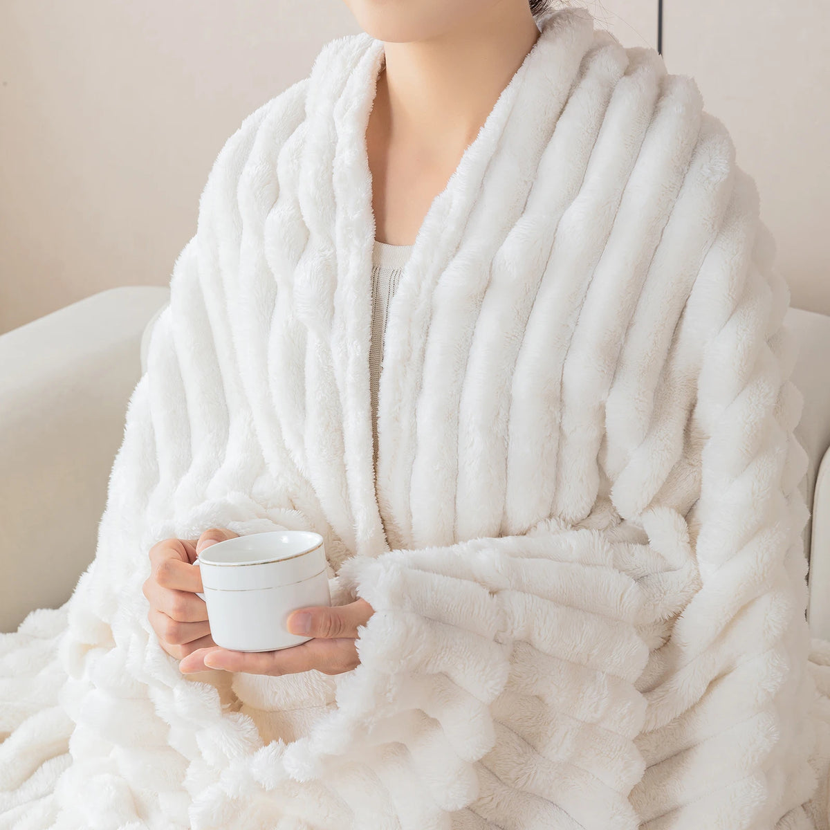 A person is wrapped in a thick white knitted blanket with vertical ribbing, holding a white mug.