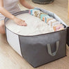 A person is placing clothing and a quilt into a gray and white portable storage bag on a wooden floor.
