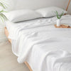 A white quilted blanket is draped over a bed, with a vase of green foliage on the side.
