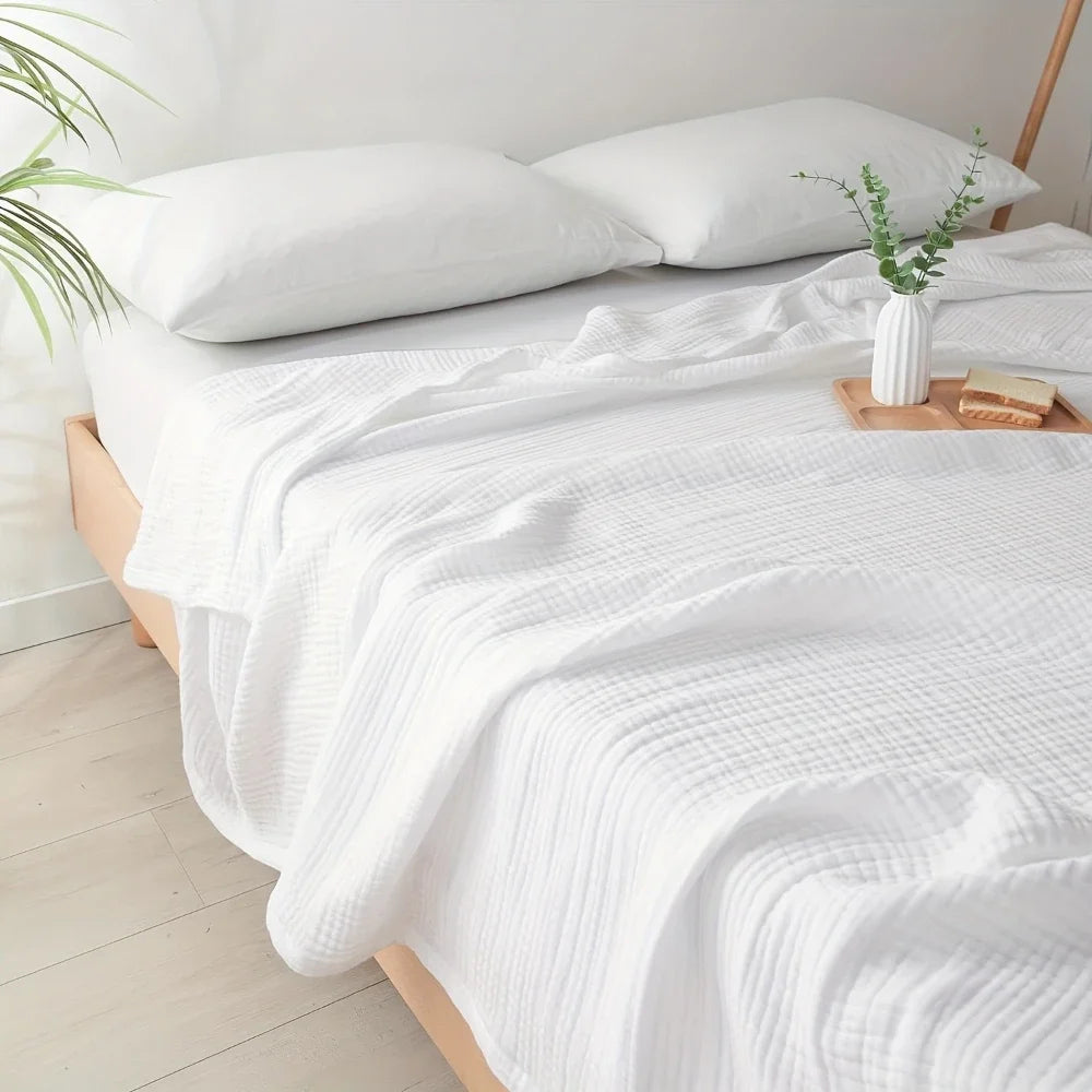 A white quilted blanket is draped over a bed, with a vase of green foliage on the side.