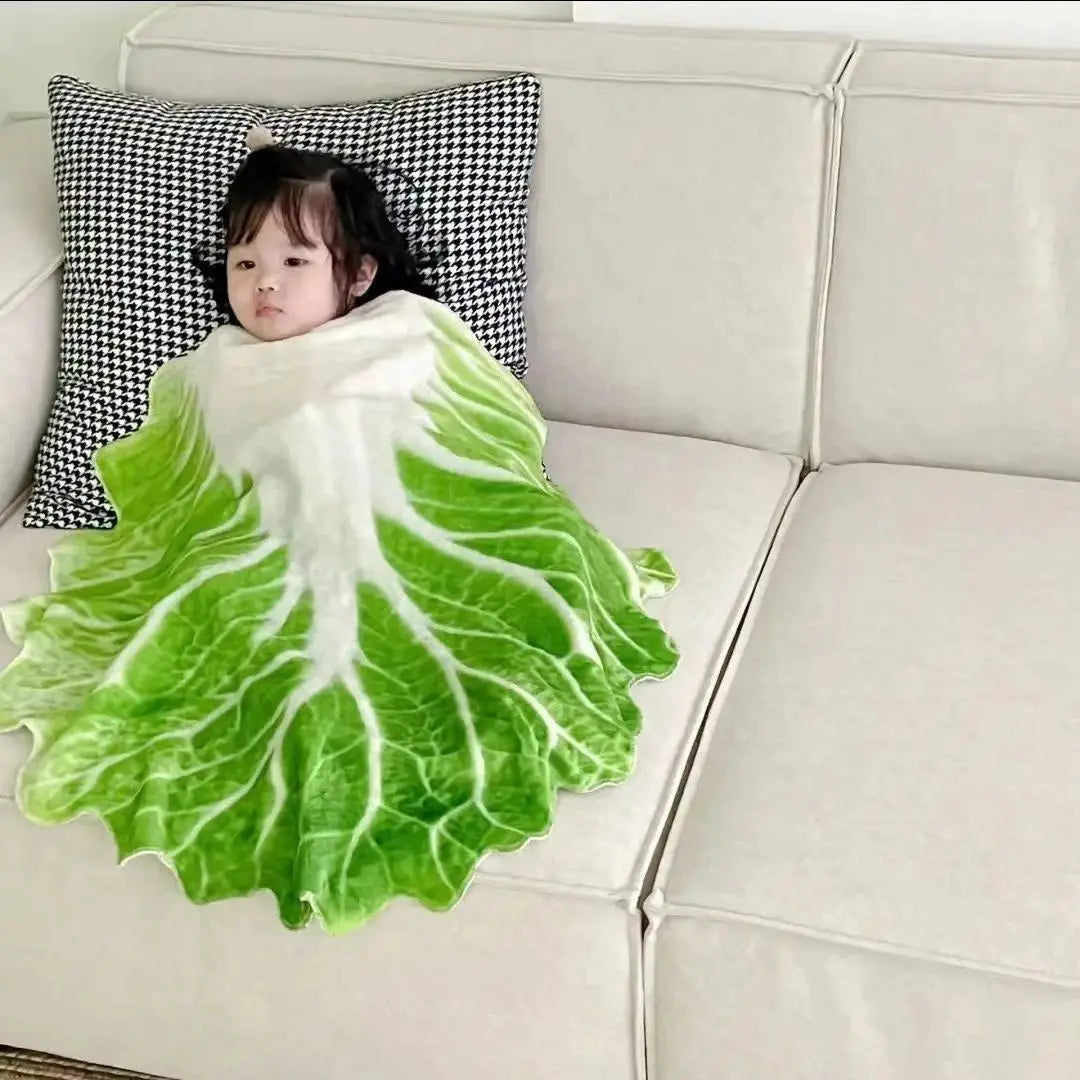 A baby is lying on a white plush throw blanket with a green leaf pattern on a light gray sofa.