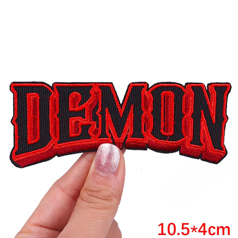 A close-up of a red embroidered patch spelling the word DEMON held by a hand.