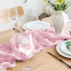 A pink cheesecloth table runner is laid on a wooden table with white plates and a glass of rosé wine.