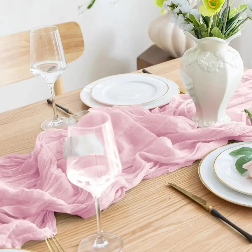 A pink cheesecloth table runner is laid on a wooden table with white plates and a glass of rosé wine.