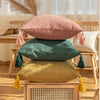 Three throw pillows in pink, green, and mustard yellow linen fabric with tasseled edges on a wooden surface.
