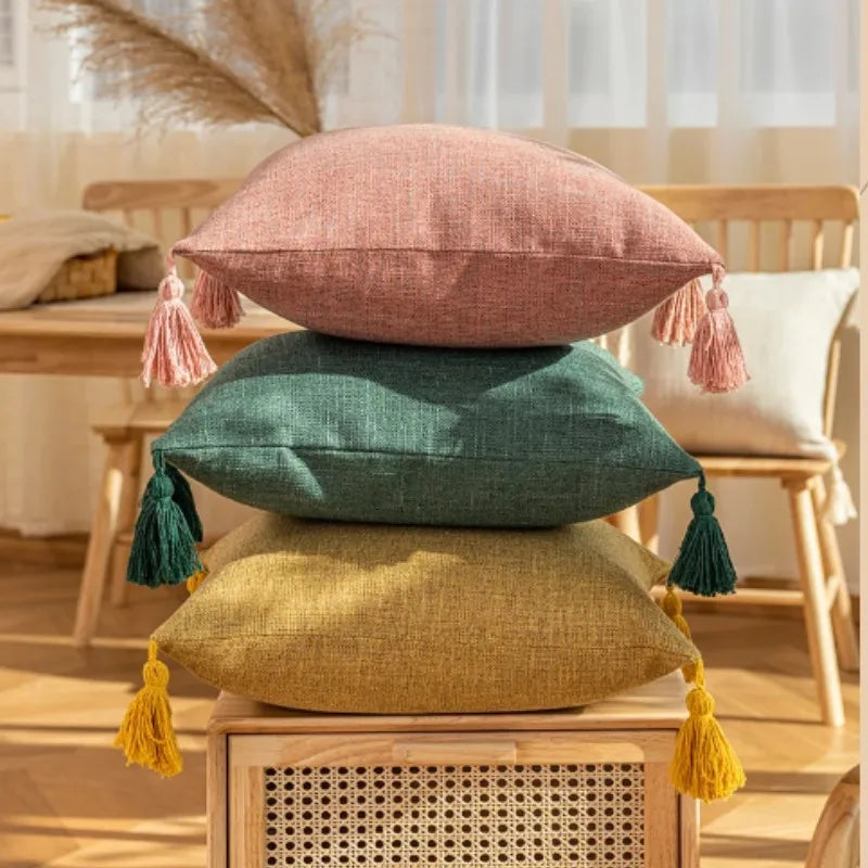 Three throw pillows in pink, green, and mustard yellow linen fabric with tasseled edges on a wooden surface.