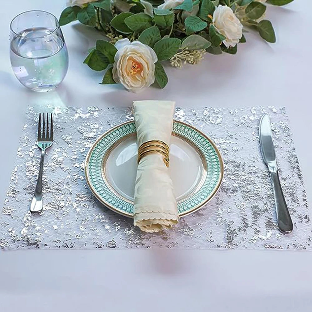 A silver-edged plate with a rolled napkin is set on a sparkly silver table runner with floral greenery and a glass of water.
