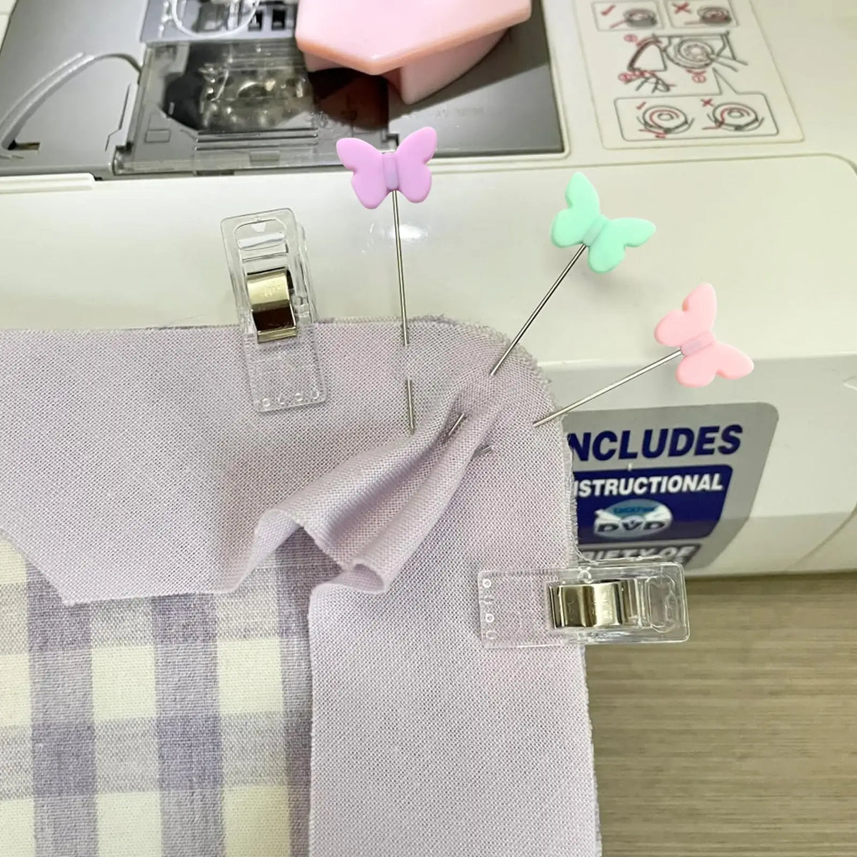 A set of colored sewing pins, including pink and green butterfly-shaped pins, displayed on a purple fabric.