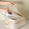 A hand opens a beige portable travel shoe organizer bag revealing a pair of white sneakers.