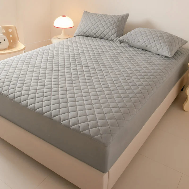 A gray quilted mattress protector is displayed on a bed with matching pillows and a bedside lamp.