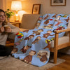 A light blue plush throw blanket featuring illustrations of C-Curious George characters on a beige sofa.