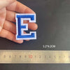 A hand holds a blue alphabet embroidery patch letter E next to a ruler showing its dimensions.