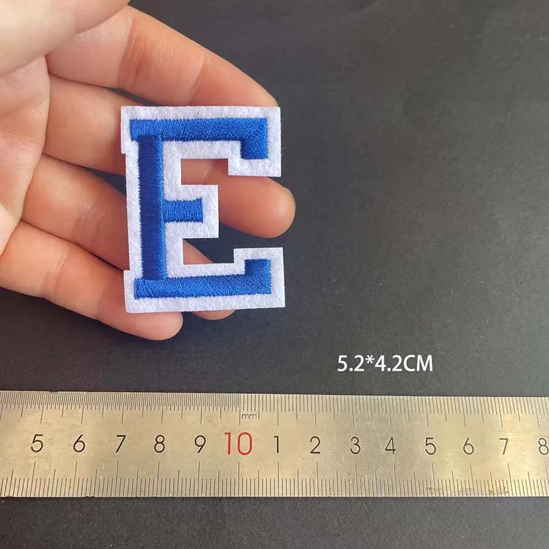 A hand holds a blue alphabet embroidery patch letter E next to a ruler showing its dimensions.