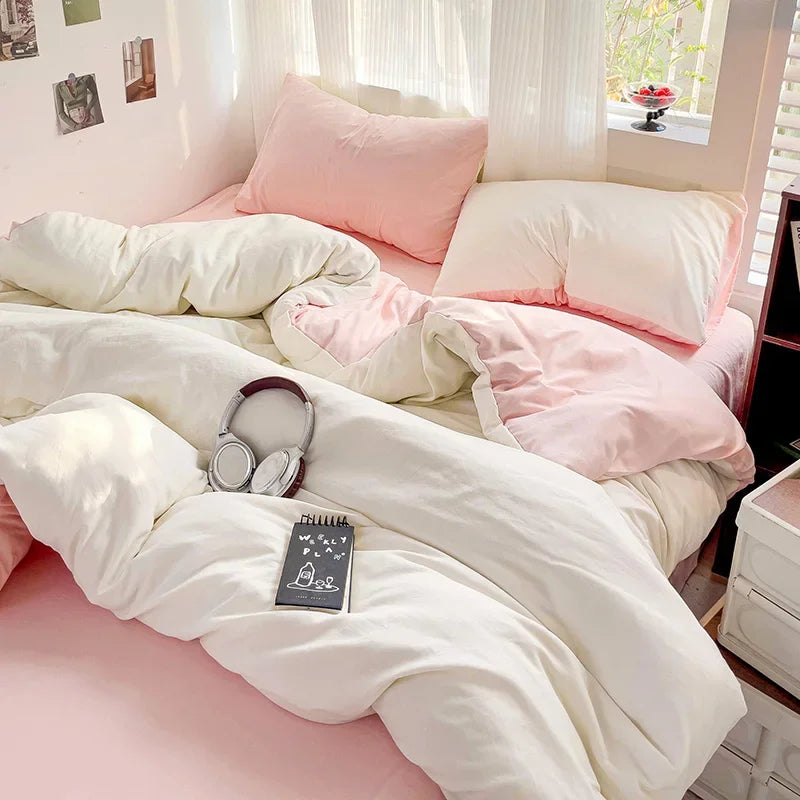 A pink and white bed with fluffy linens, including pillows and a duvet, next to a window.