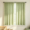 A pair of light green floral print curtains hanging in a window with white trim and a wooden furniture piece nearby.