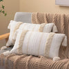 A beige knitted throw pillow cover with cream accents and tassels resting on a textured brown sofa.