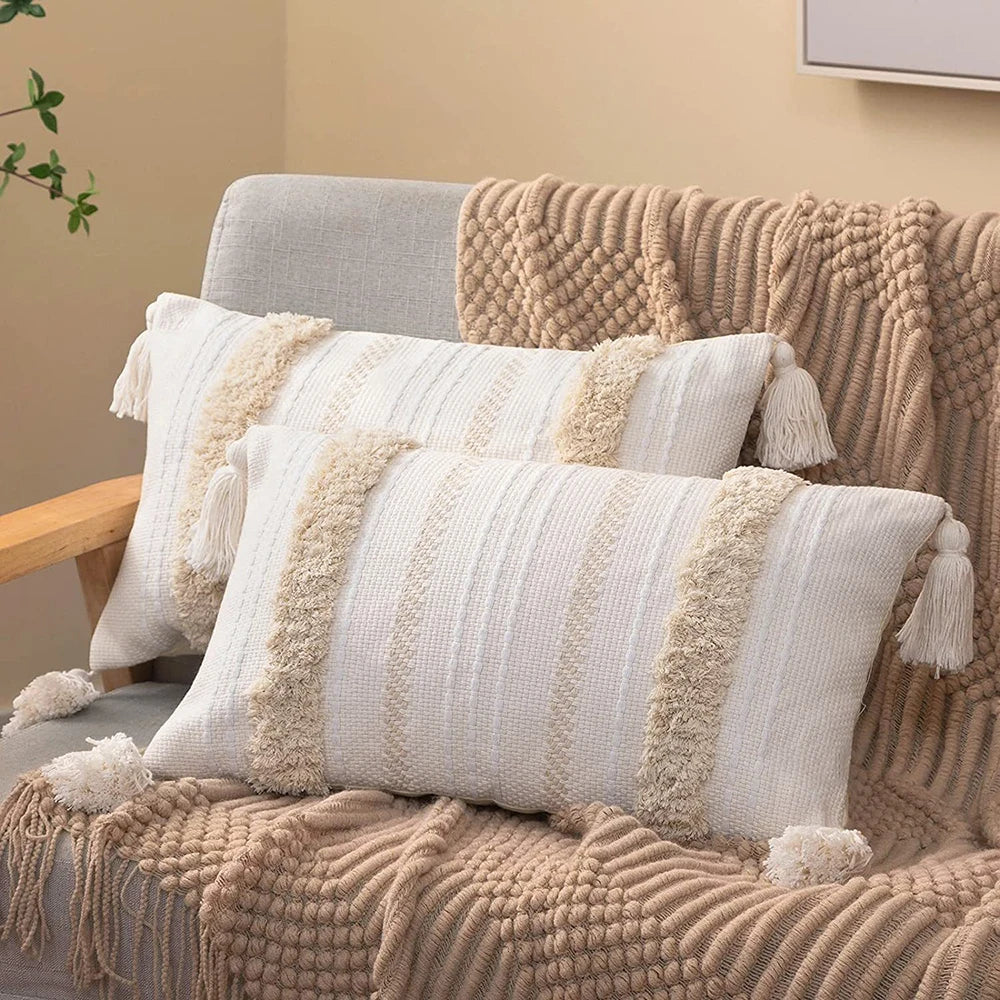 A beige knitted throw pillow cover with cream accents and tassels resting on a textured brown sofa.