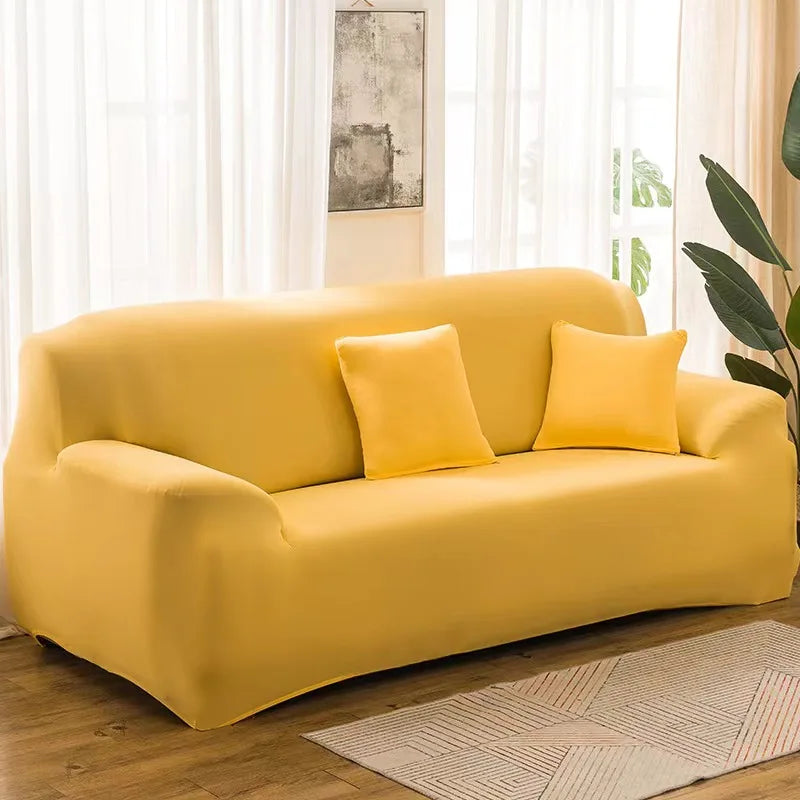 A mustard yellow stretch sofa cover with two accent pillows placed on a wooden floor near a window.