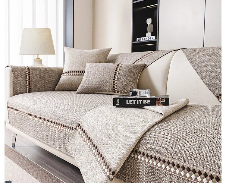 A beige linen sofa cover with decorative piping and cushions displayed in a bright living room setting.