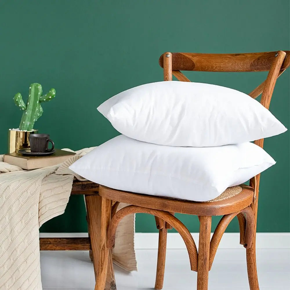 A white cotton-filled decorative throw pillow insert resting on a wooden chair against a dark green wall.