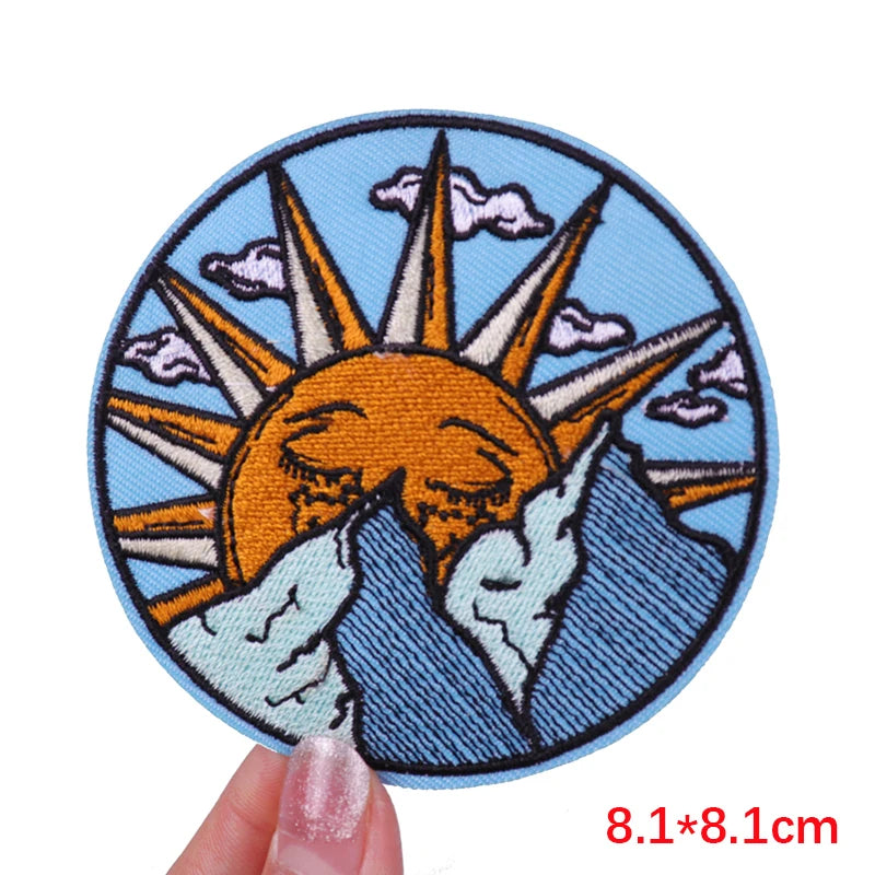 An embroidered patch featuring a stylized orange sun rising over blue mountains and clouds.