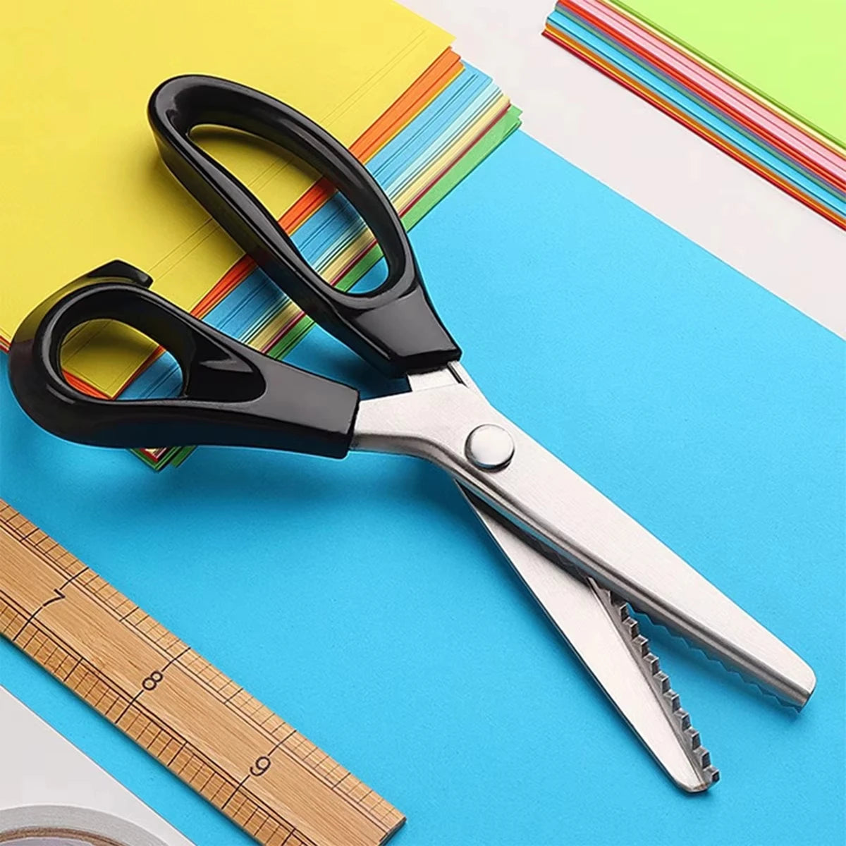 A pair of black serrated edge fabric scissors with stainless steel blades resting on a blue background next to colorful paper