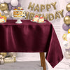 A deep burgundy satin tablecloth is set for a birthday celebration with decorated cakes and gold balloons.