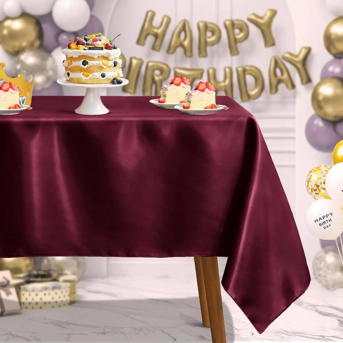 A deep burgundy satin tablecloth is set for a birthday celebration with decorated cakes and gold balloons.