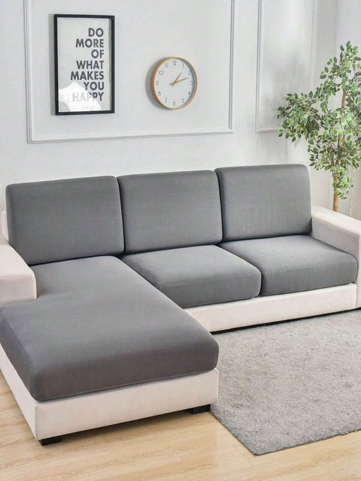 A modern gray sofa with slipcovers sits in a bright living room next to a framed motivational sign and a potted plant.