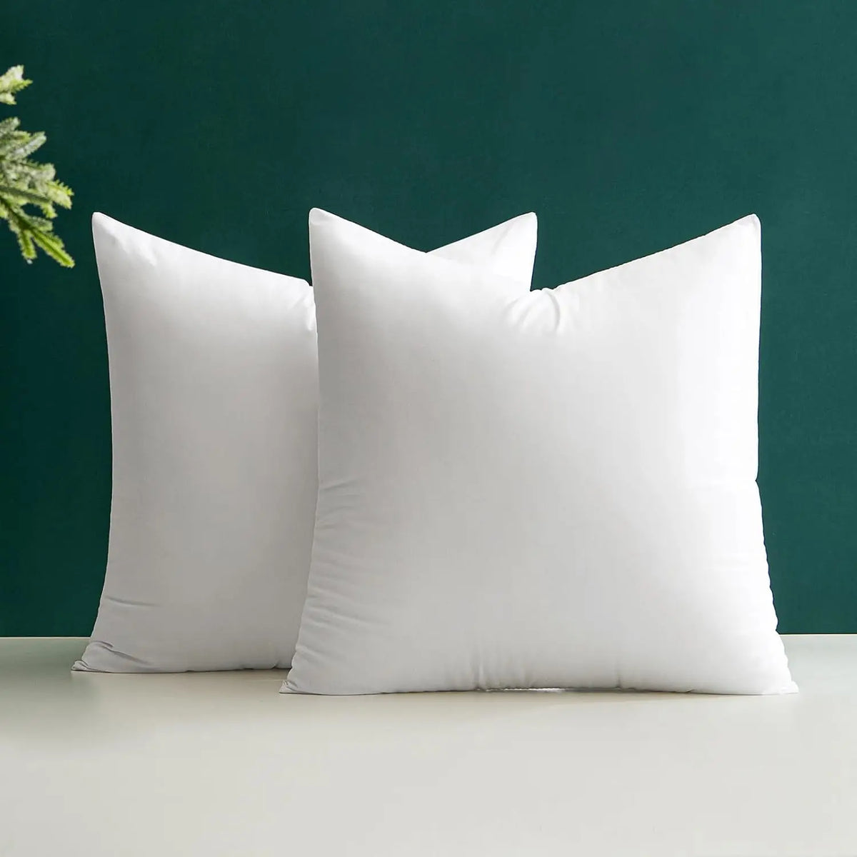 A pair of white polyester cushion inserts for throw pillows resting on a light surface against a dark green background.
