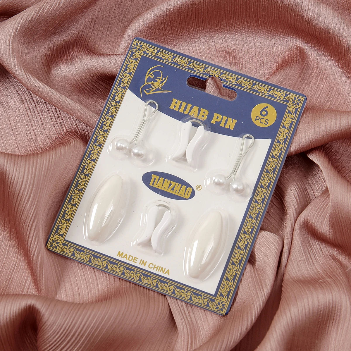 A package of six plastic safety pins for hijab accessories is displayed on pink fabric.