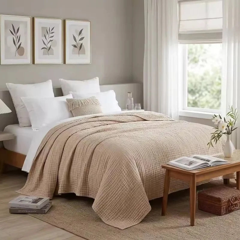 A beige pure cotton gauze throw blanket draped over a white bed with framed art on the wall.