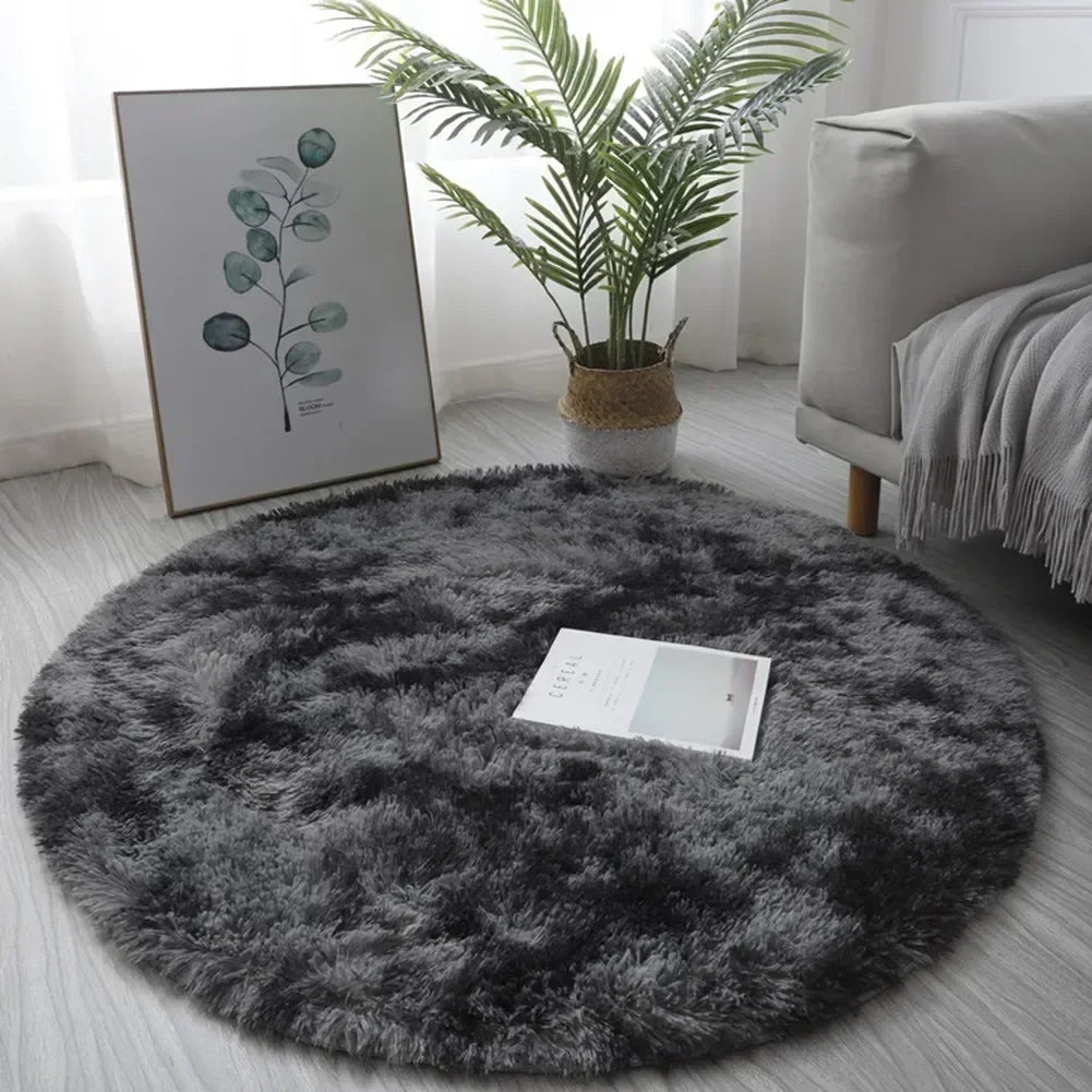 A round, dark gray shaggy area rug placed on a light wooden floor next to a potted palm plant and framed artwork.
