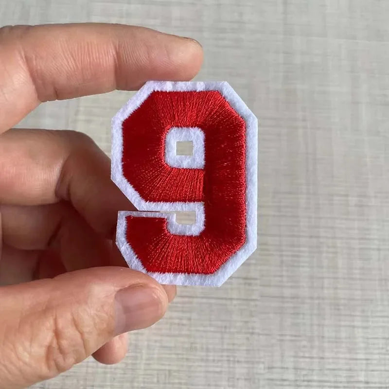 A hand holds a red and white embroidered number 9 patch for apparel.