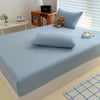 A light blue fitted bed sheet cover is placed on a bed, with pillows and a teddy bear visible.