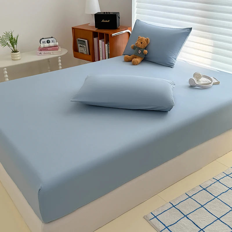 A light blue fitted bed sheet cover is placed on a bed, with pillows and a teddy bear visible.