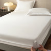 A white fitted sheet is laid on a bed, with pillows and a bedside lamp visible.