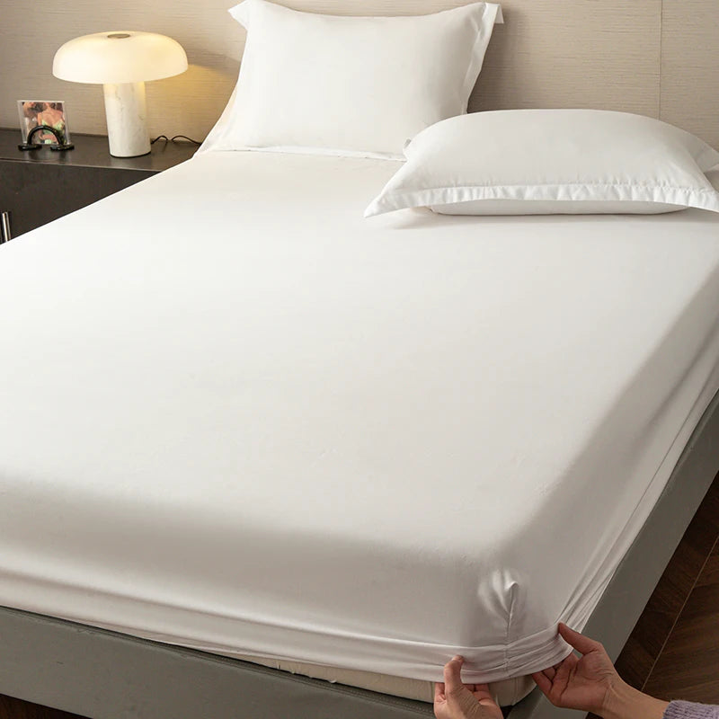A white fitted sheet is laid on a bed, with pillows and a bedside lamp visible.