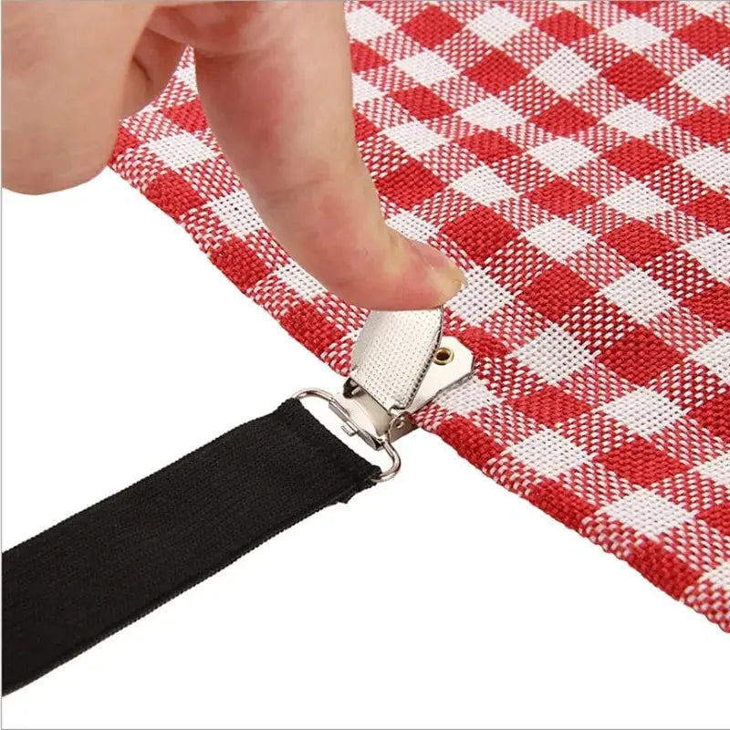 A hand is holding a black strap attached to a red and white checkered bed sheet with silver clips.