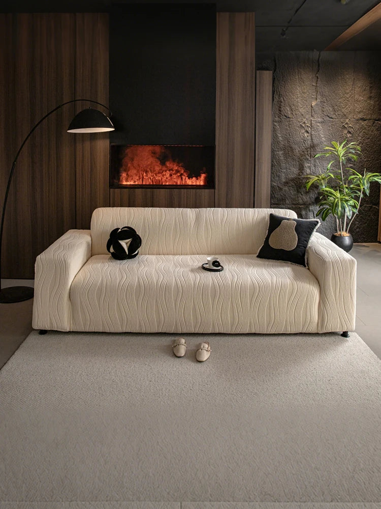 A cream-colored stretchable sofa cover with a vertical line pattern sits on a light rug in a modern, dark-toned room.