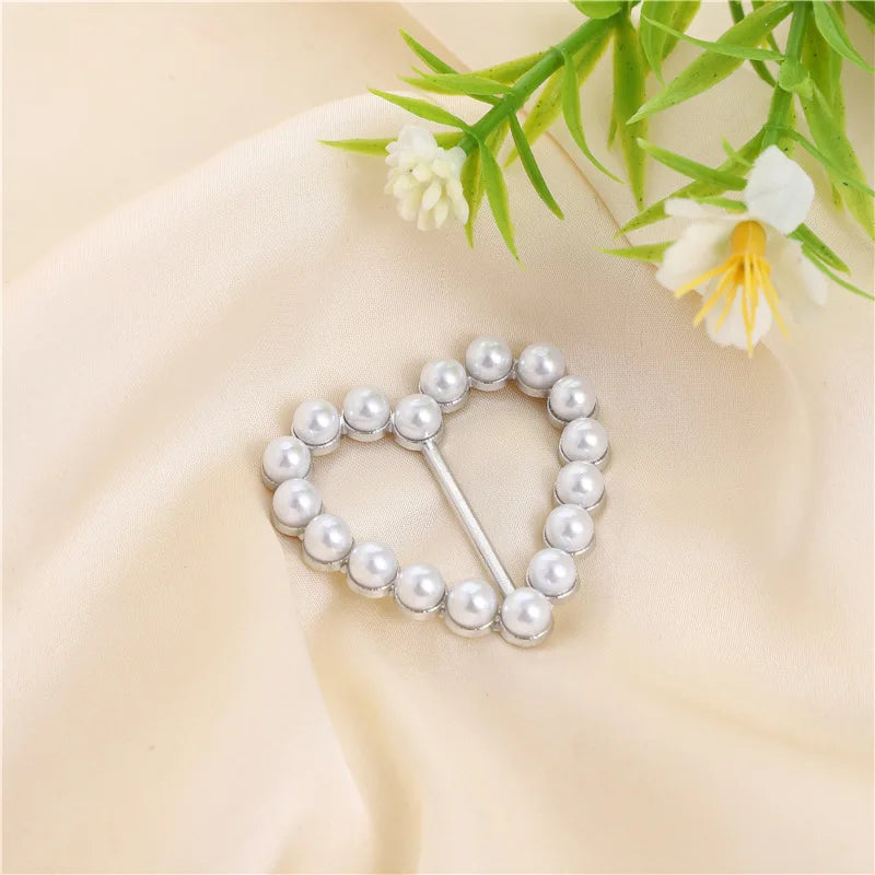 A delicate silver heart-shaped ring clip made of small pearls rests on soft beige fabric with green foliage.