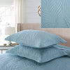 A set of light blue embroidered cotton quilted pillow covers displayed on a bed with a white window and light bedding.