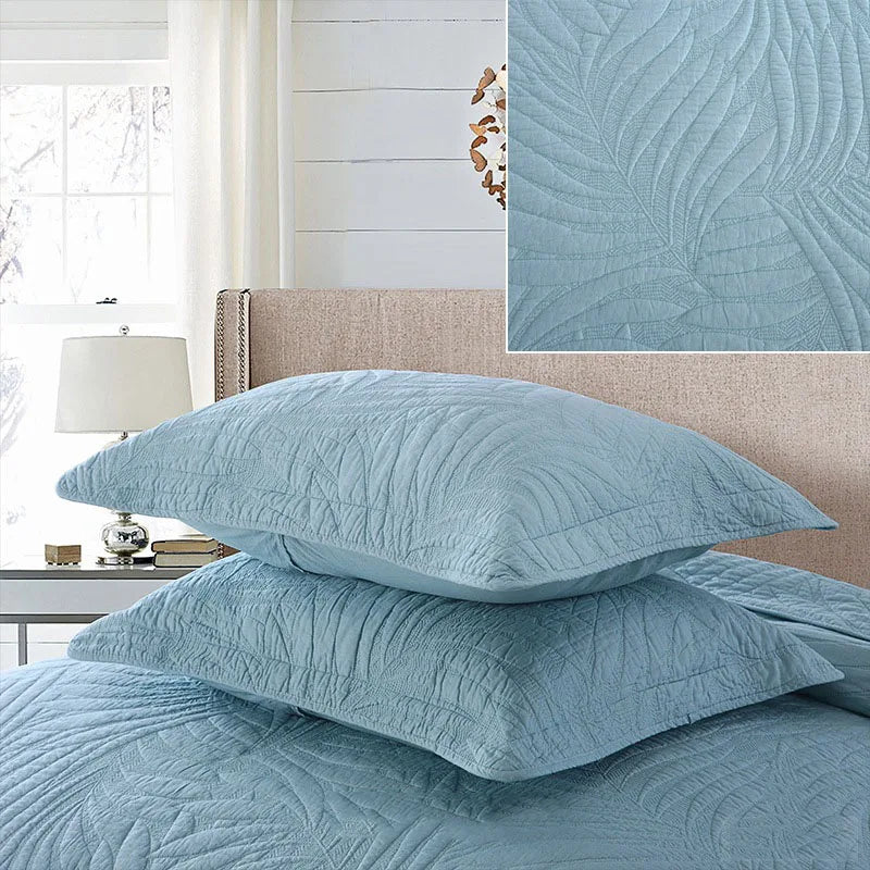 A set of light blue embroidered cotton quilted pillow covers displayed on a bed with a white window and light bedding.