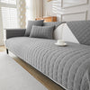 A gray quilted anti-slip sofa cover with white accents and pillows displayed on a modern sofa.